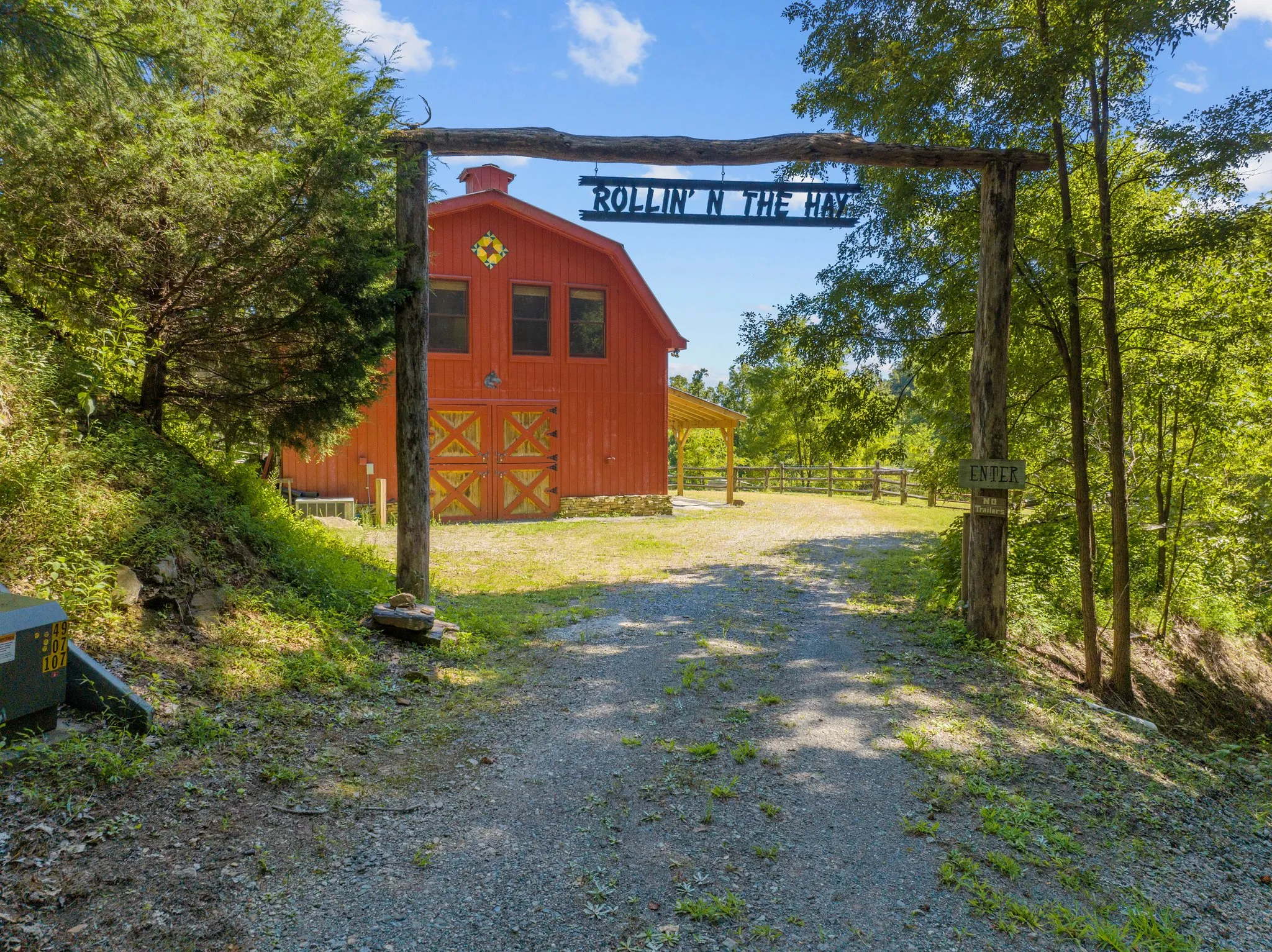 Red gambrel barn with "Rollin' N The Hay" sign above entrance archway surrounded by forest